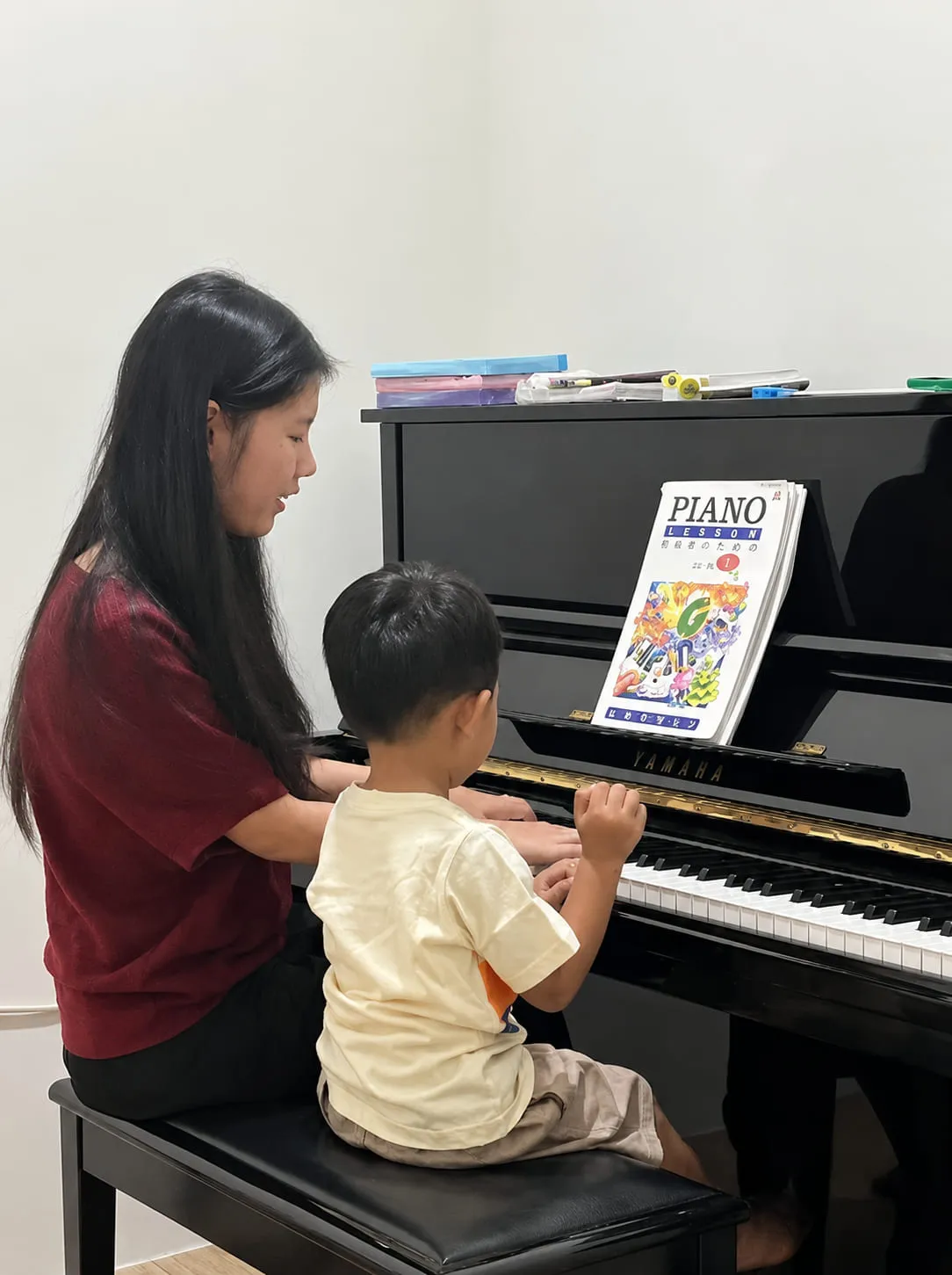 Pengajar Cendana mendampingi seorang anak bermain piano, dengan buku not terbuka di depan mereka