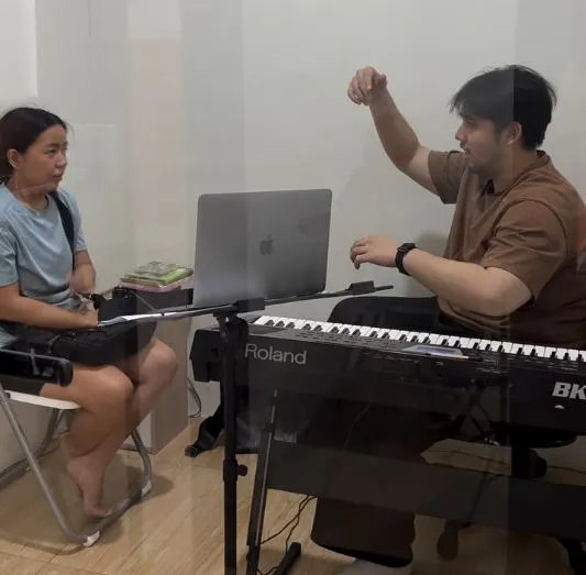 Pengajar mendampingi murid dewasa berlatih vokal sambil membaca not di keyboard