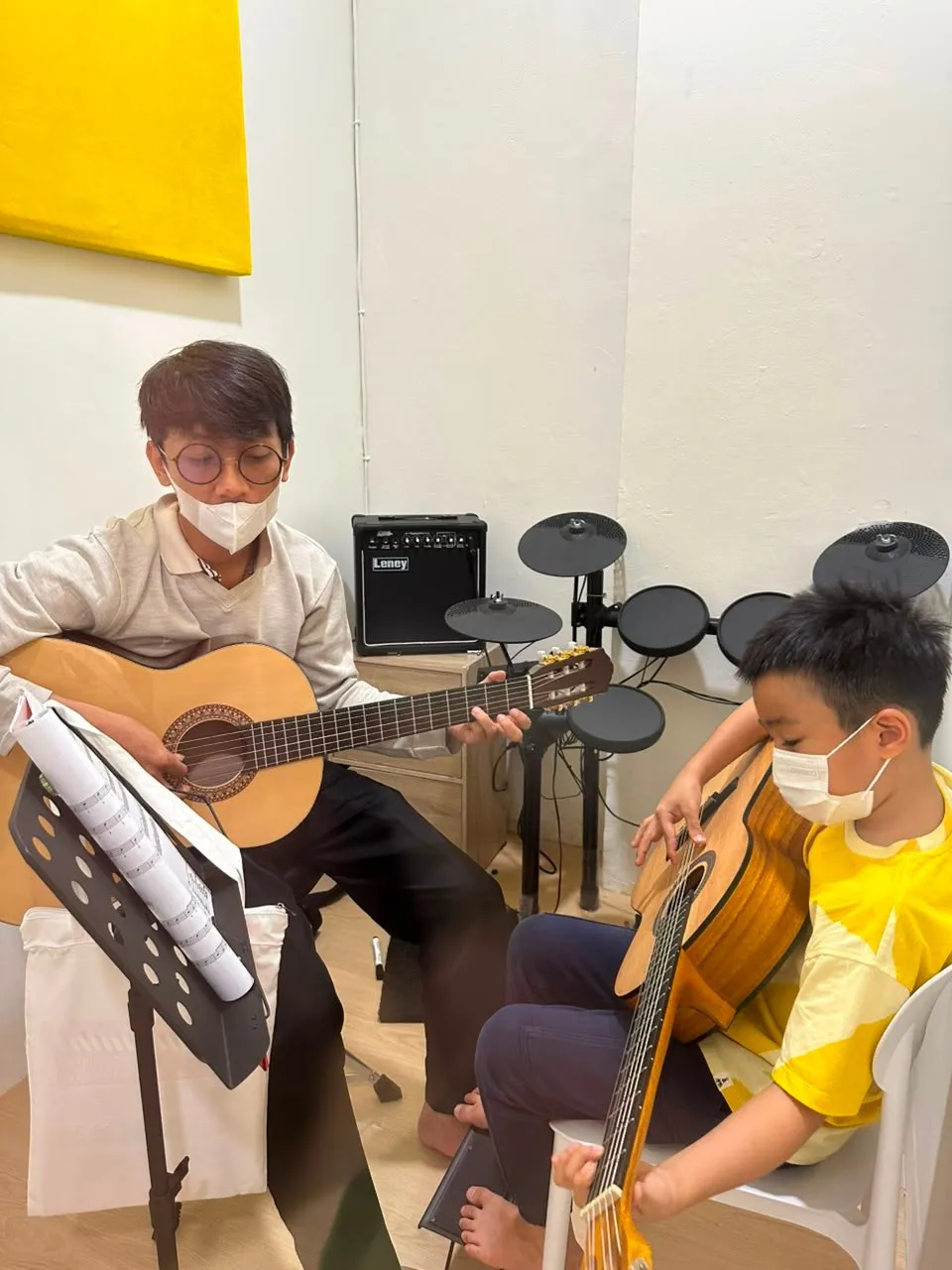 Pengajar mendampingi murid berlatih gitar, dengan drum elektrik di ruang yang sama